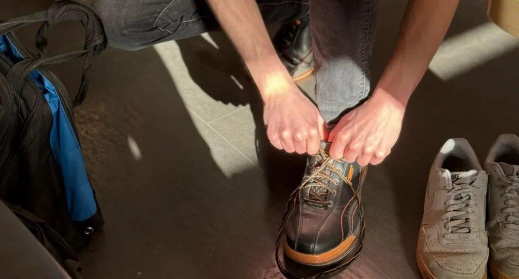 Person bindet Bowling-Schuhe vor dem Spiel, Sneaker und Sporttasche stehen daneben auf dem Boden